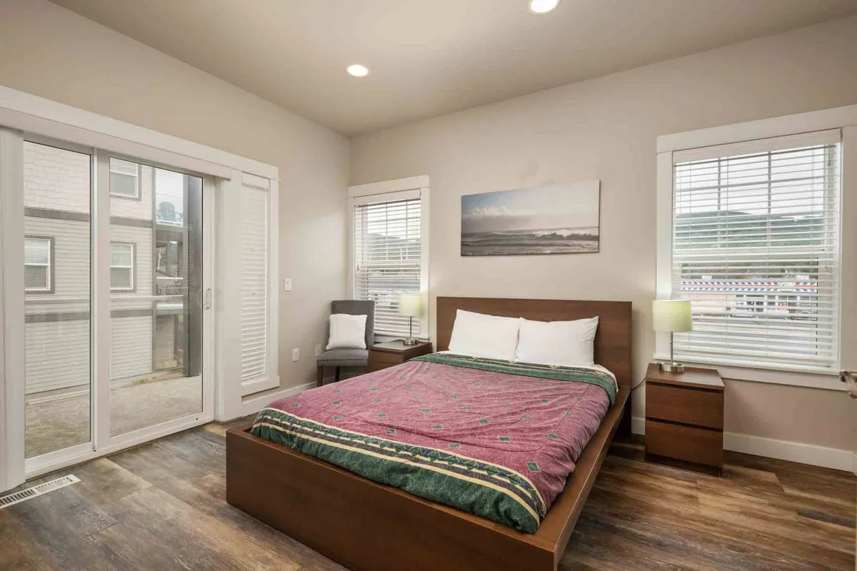 A bright bedroom boasting a queen bed with colorful bedding, large windows letting in warming natural light, artwork on the wall, and nightstands with lamps for added comfort. The room has a fresh, serene feel that invites relaxation.