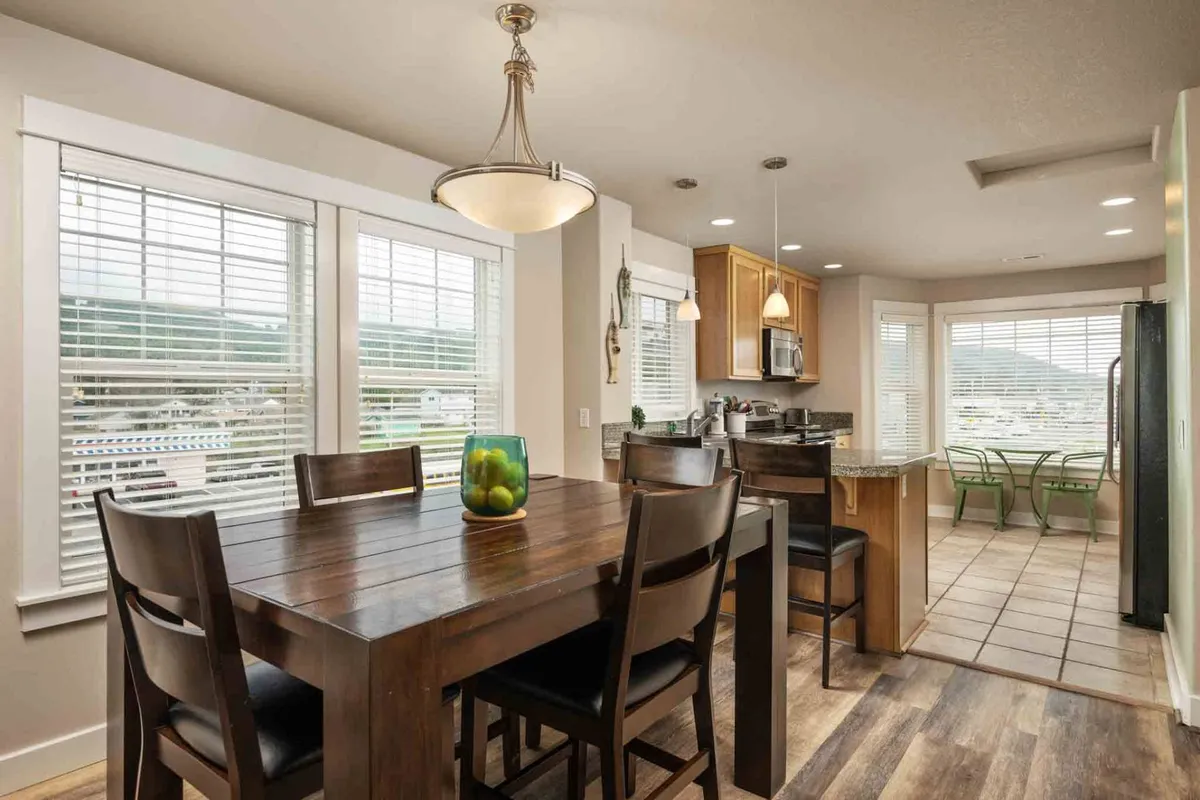 A bright and welcoming dining area with a large table, green chairs, a kitchen counter in the background, and huge windows that let in plenty of sunlight. The scene conveys a sense of togetherness and everyday joy.