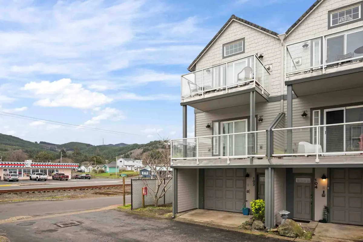 An exterior view of the townhouse showcases multiple balconies, garage doors, and the surrounding landscaped area. The design suggests an appealing blend of privacy and community, with balconies clearly visible as essential outdoor extensions of the home. The scene captures the welcoming ambiance of a beachside townhouse complex steps from local attractions and the shoreline.