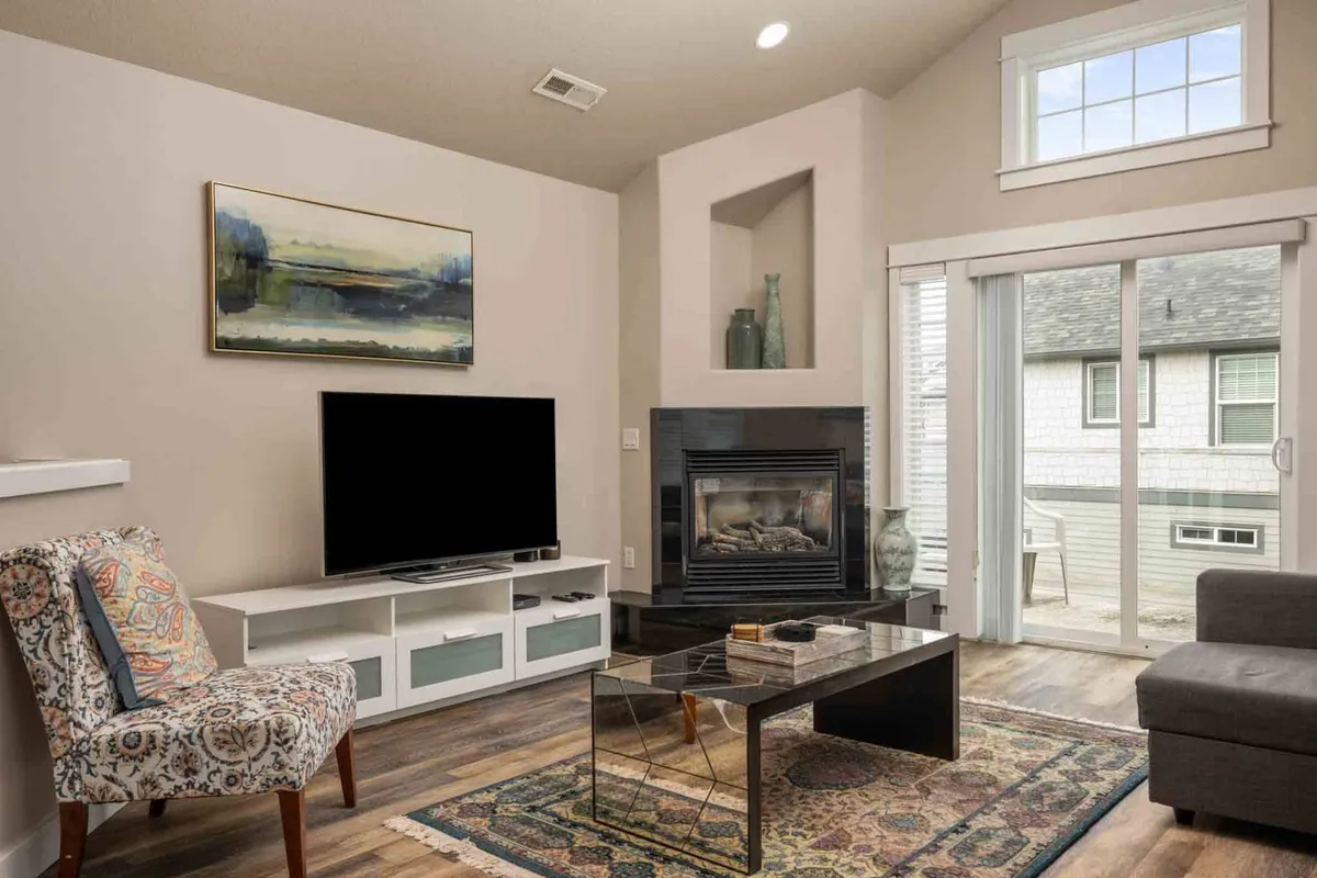 A welcoming living area featuring a cozy armchair, fireplace, coffee table, and artwork. Vaulted ceilings and generous windows fill the space with natural light, creating an inviting environment for both relaxation and productivity. The arrangement encourages reading, casual work, or unwinding after a fulfilling day by the waves.