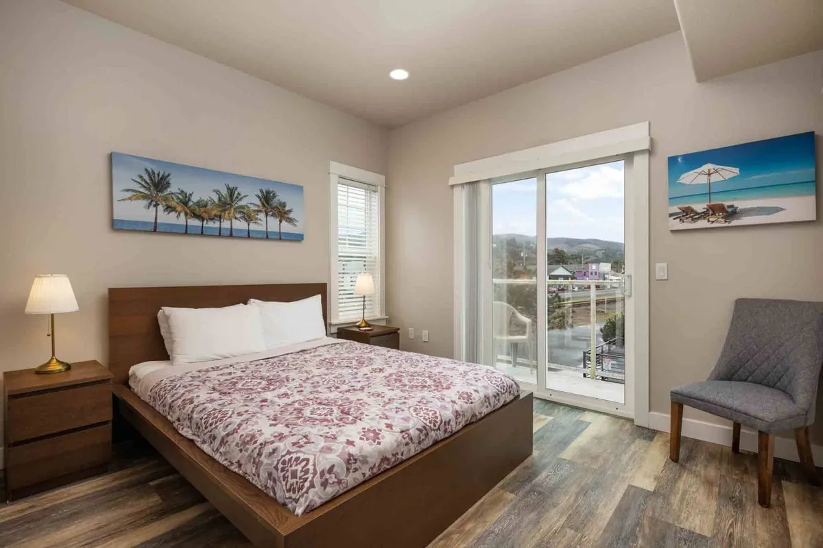 A peaceful bedroom featuring a queen-size bed with soft bedding, tropical-themed artwork on the walls, a comfortable chair, a lamp, and a sliding glass door that lets in abundant natural light. The décor utilizes gentle coastal hues to evoke a relaxing ambiance.