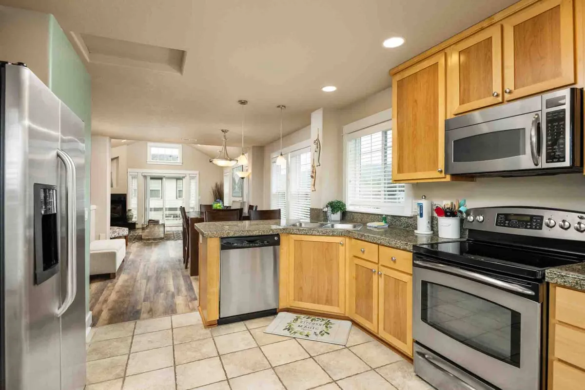 An inviting kitchen space with stainless steel appliances, granite countertops, warm wooden cabinetry, and a dining area visible in the background. Large windows bring in ample natural light, creating a cheerful and practical atmosphere.