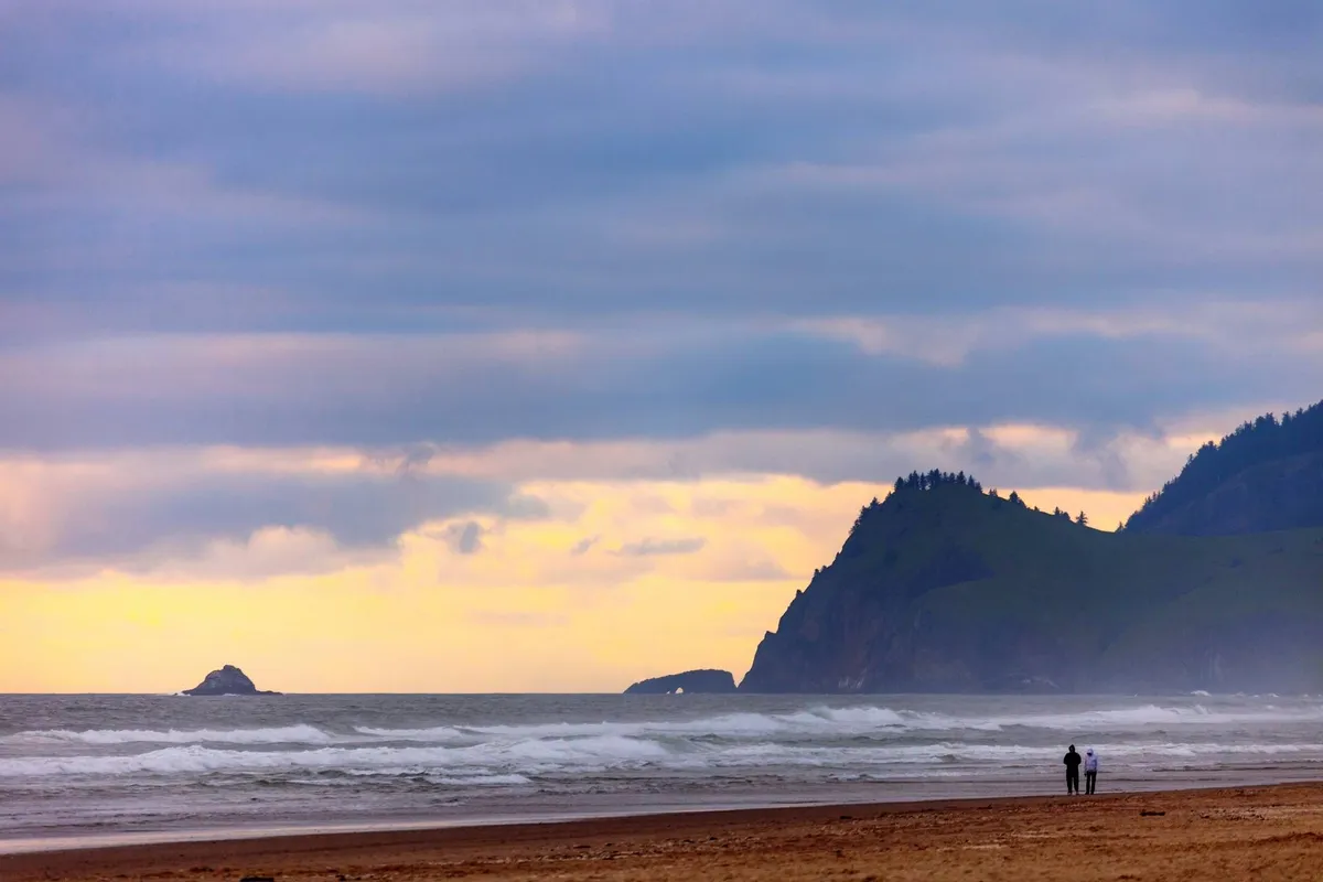 A picturesque stretch of Rockaway Beach at sunset, with gentle waves rolling onto the sand. In the foreground, a couple walks hand-in-hand along the shoreline. Distant cliffs and a unique rock formation frame the scene, capturing the area's natural beauty and romantic atmosphere.