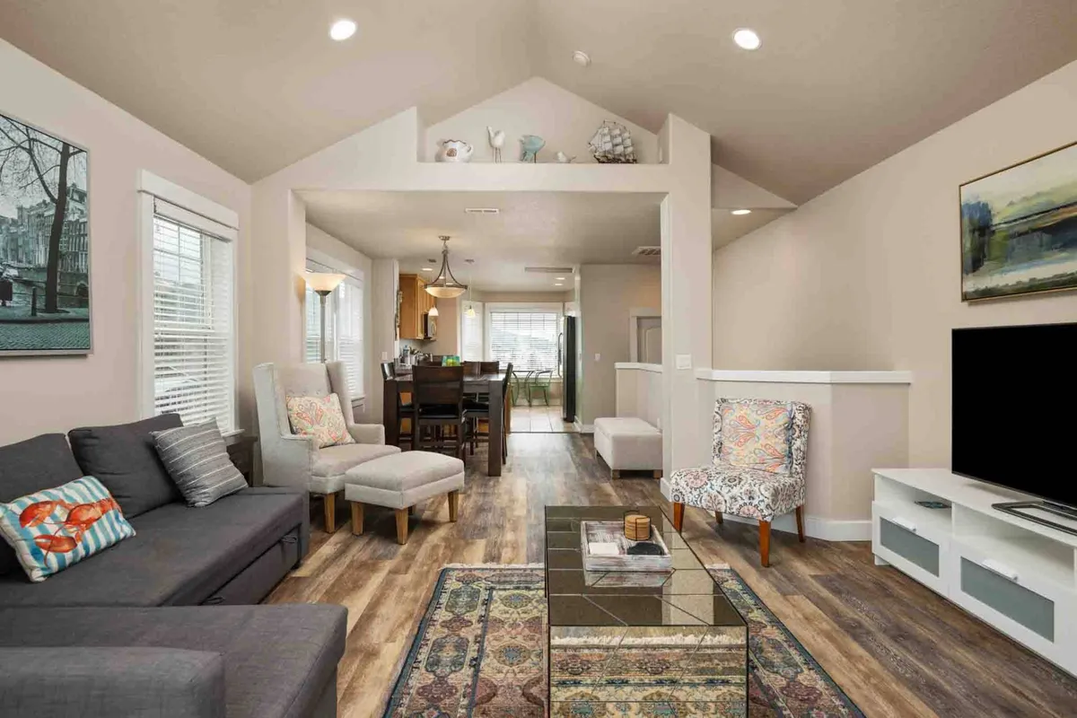 The image showcases a cozy, light-filled living area featuring a plush sofa with vibrant, colorful cushions, a modern armchair, a sleek glass coffee table, and tasteful artwork on the wall. The inviting space is perfect for relaxing, entertaining, or enjoying quiet moments with family and friends.
