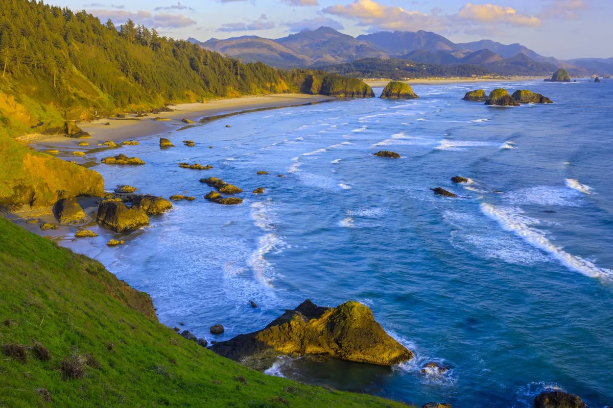 This image showcases a panoramic view of the Oregon Coast, featuring rolling ocean waves, rugged rock formations, distant mountains, and a sandy shoreline. The landscape is tranquil, inviting beachgoers to explore, play, and soak in the coastal scenery.