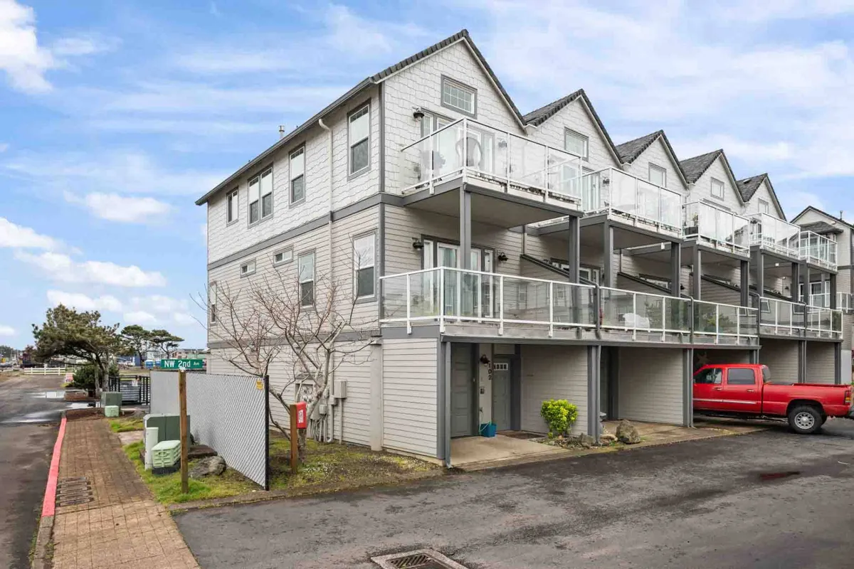 The building's modern exterior with balconies featuring glass railings. A red pickup truck is parked out front, and residents can be seen relaxing and socializing on the balconies. The scene conveys a welcoming, active community atmosphere with the promise of ocean breezes and sunset views.