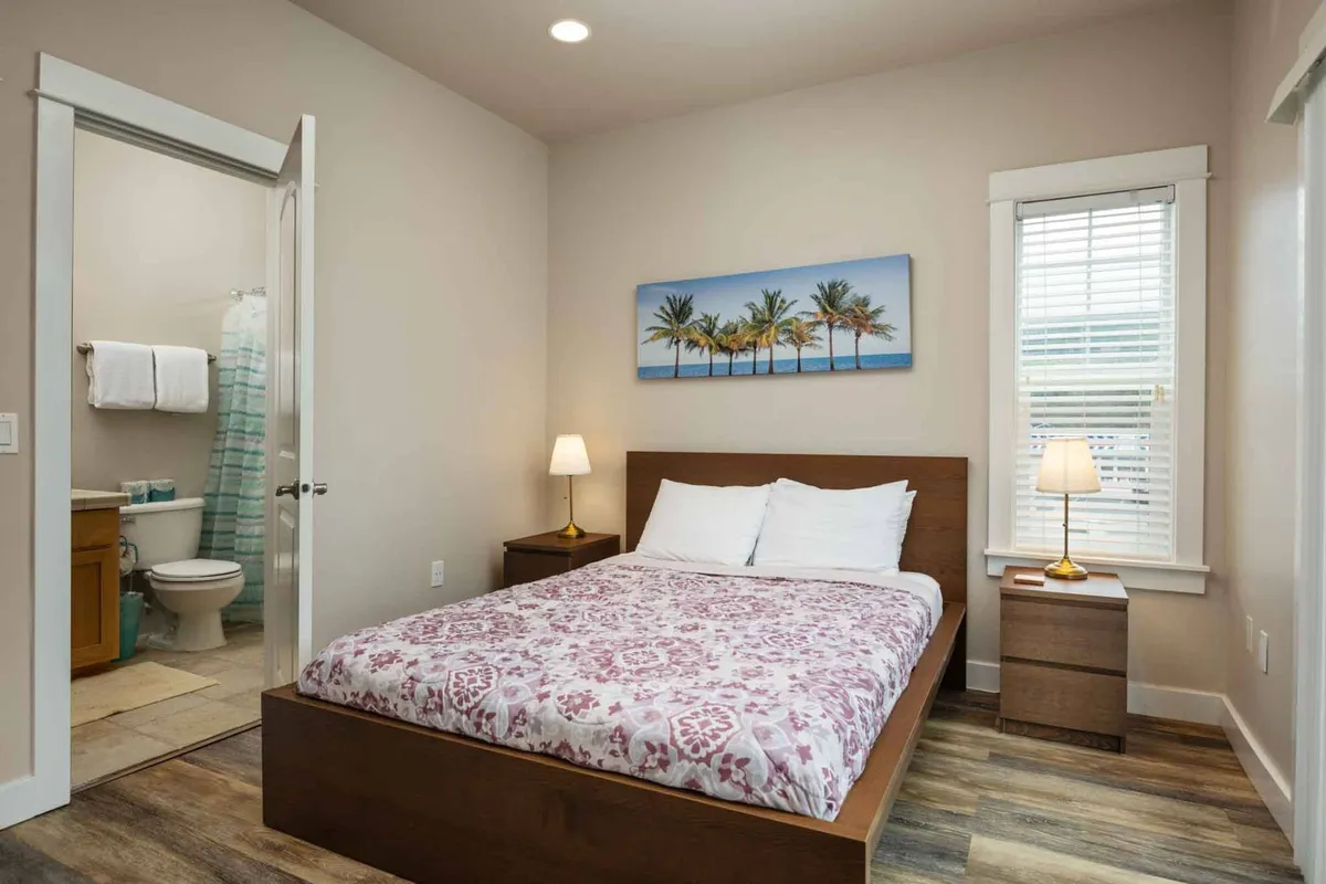 A cozy bedroom with a queen bed adorned in patterned bedding, a framed beach scene above the headboard, a table lamp providing soft glow, and a nearby bathroom doorway. Natural light streams in, creating a calm, inviting atmosphere.