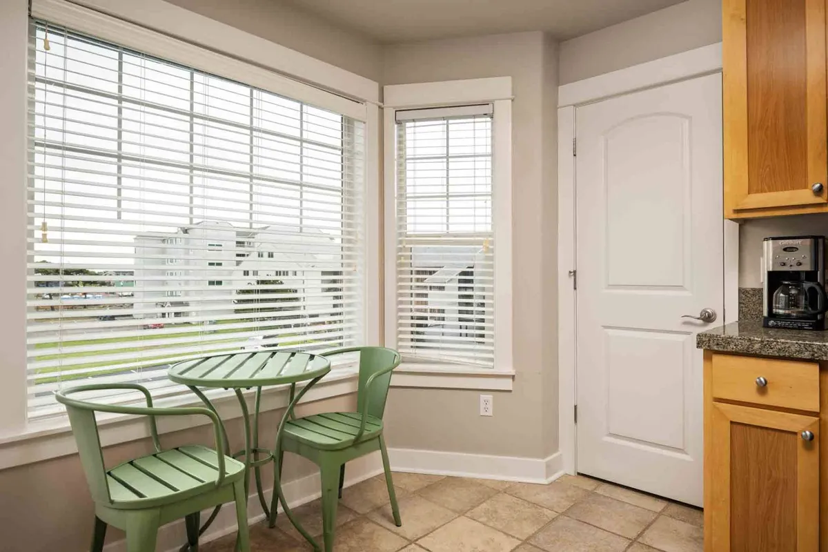 A cozy dining nook with green chairs and a round table, framed by large windows that let in ample natural light. The setting is intimate yet casual, ideal for conversation, small meals, or planning the day's adventures with friends.